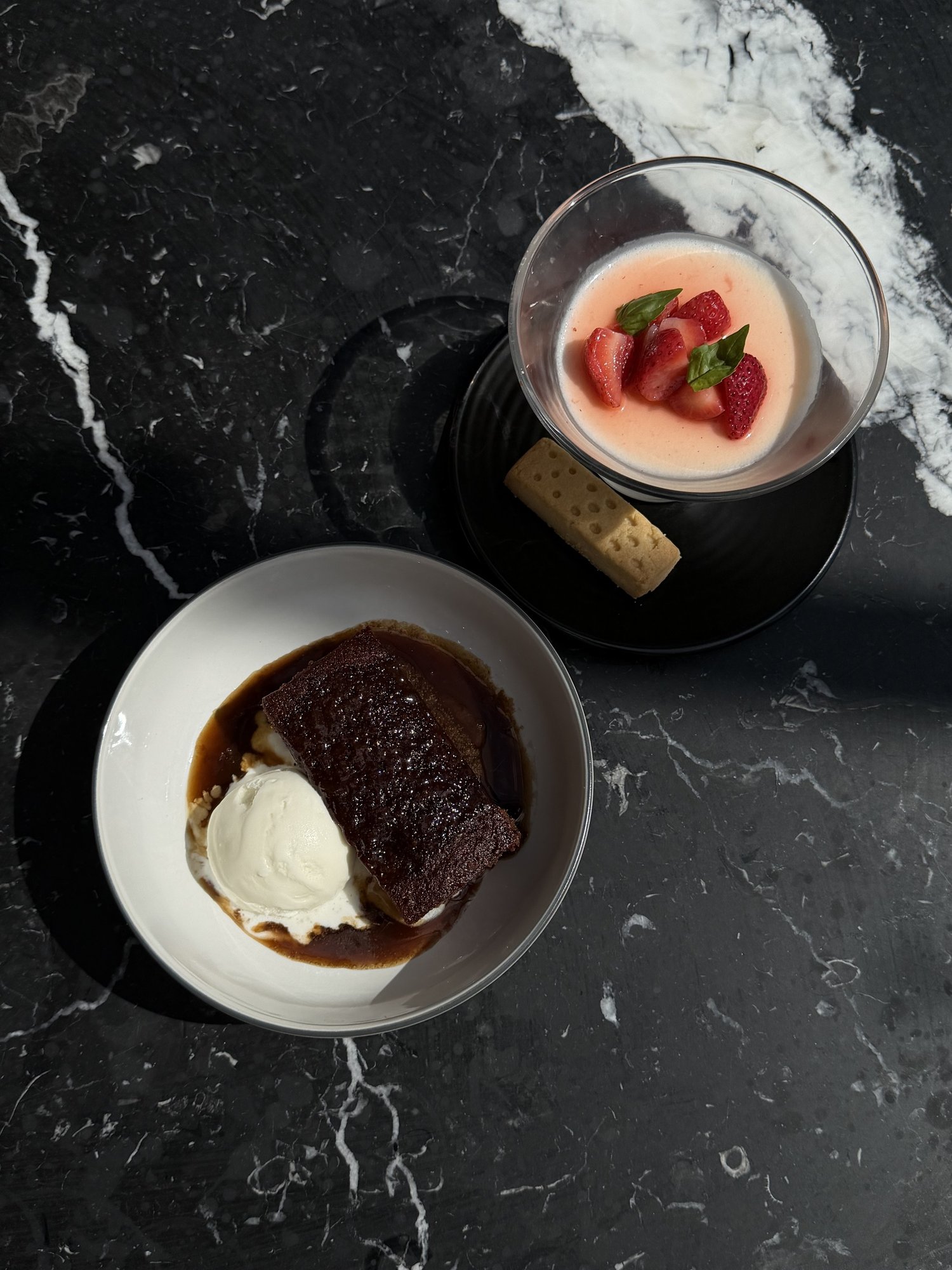 Overhead of sticky toffee pudding with ice cream beside a vanilla panna cotta topped with strawberries and basil