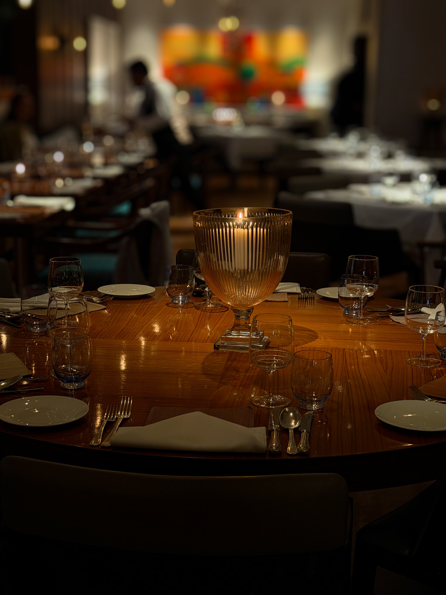 A round table laid with white napkins and glassware around a ribbed glass candle holder, the canvas lit beyond