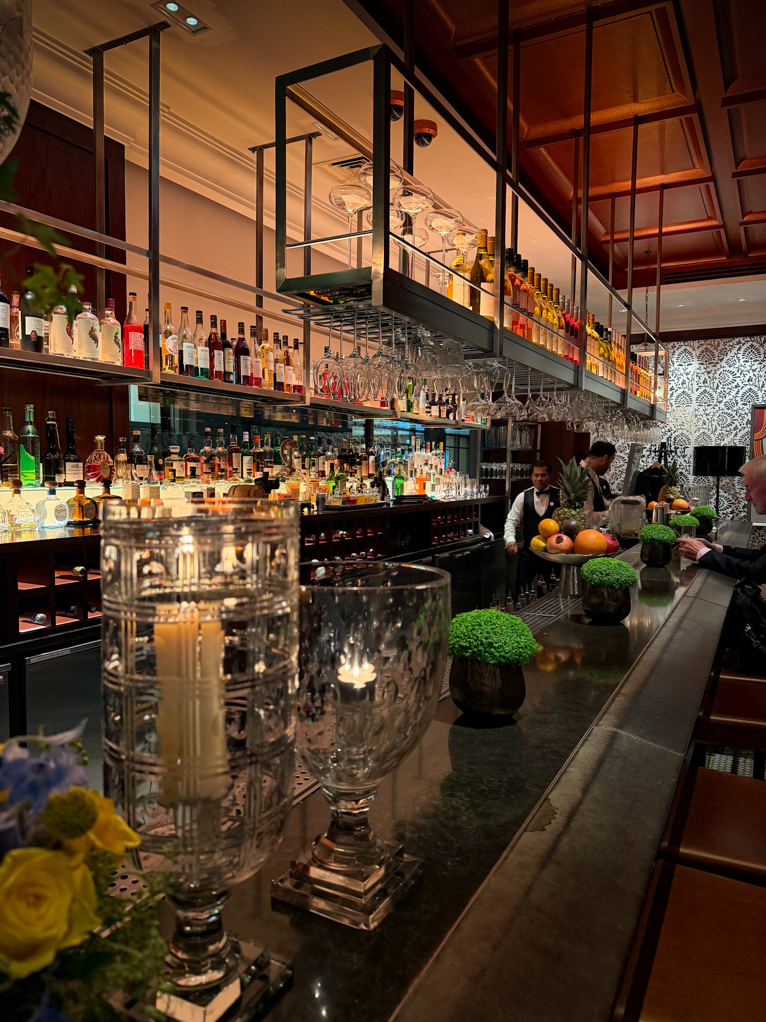 The Pukka Bar at Chutney Mary, bottles stacked beneath a suspended brass rack of glassware with bowls of topiary and citrus along the counter