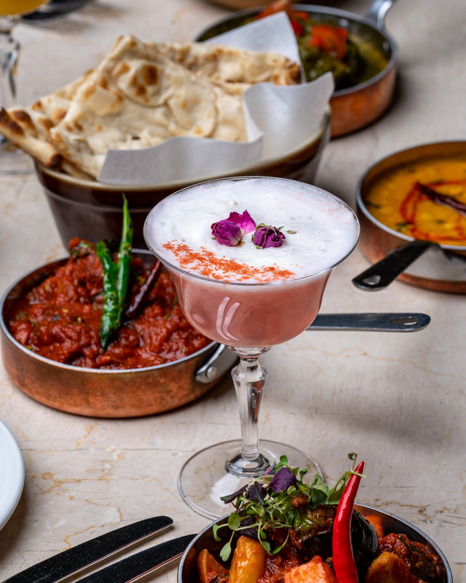 A pink cocktail topped with dried rose petals beside copper pans of curry, dal, naan and grilled dishes on a marble table
