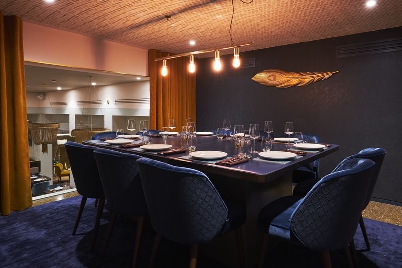 The Peacock Room at Kahani, a long table with blue velvet chairs, gold curtains and a gilded peacock feather on the wall