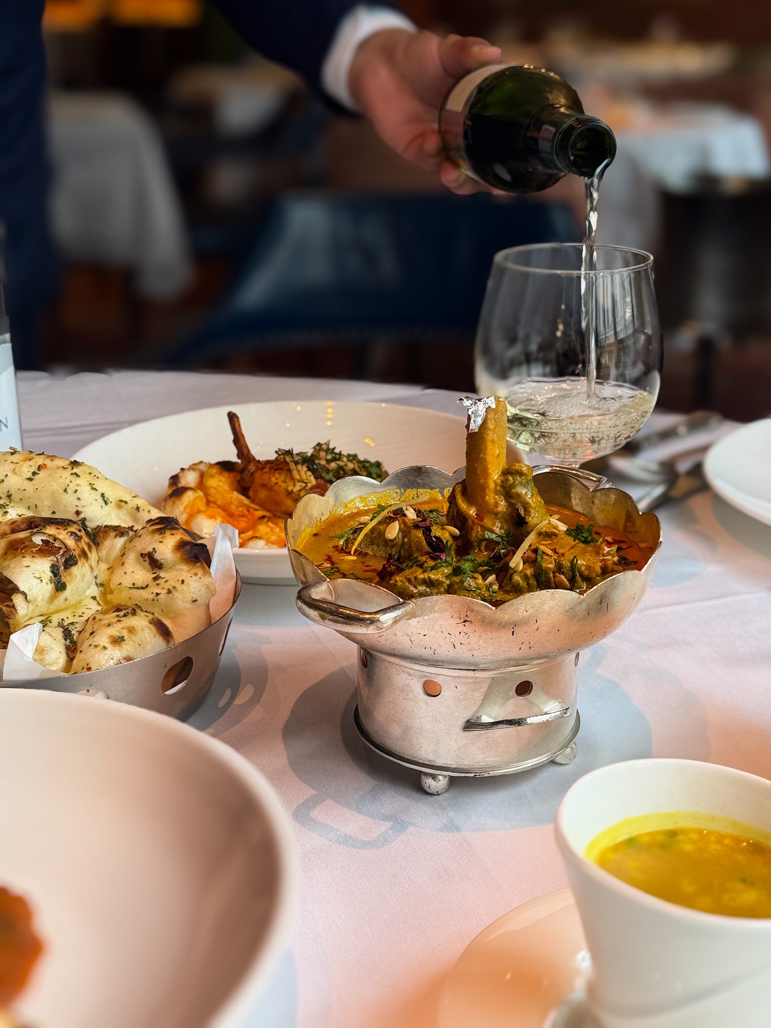 White wine being poured at the table, with naan and the lamb salan in its silver bowl